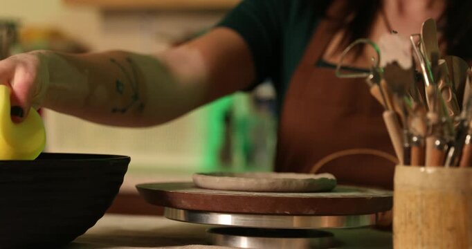 A close-up of a potter's hands using a damp yellow sponge to smooth and refine the edges of a raw clay disk on a rotating pottery wheel.

