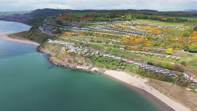 Aerial cinematic drone tracking shot of a passenger train traveling along the scenic coastline of Pettycur Bay with a caravan park on the hillside, Fife, Scotland.