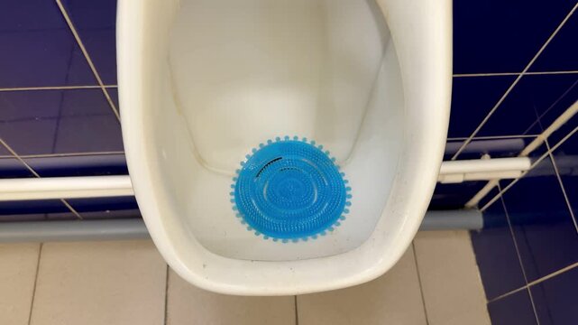 Public men's bathroom white urinal, set against glossy blue and yellow tiles, chrome flush pipe, blue deodorizer block in bowl