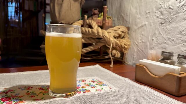 Close-up glass hazy beer on embroidered tablecloth, dim rustic restaurant interior with nautical rope and burlap sack