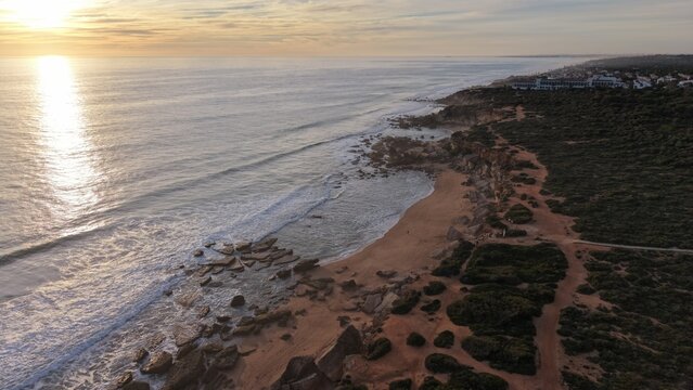 Aerial view of the Calas de Roche cliffs at sunset in Conil, Spain. Cinematic drone shot of the red sandstone cliffs and turquoise Atlantic Ocean at golden hour.