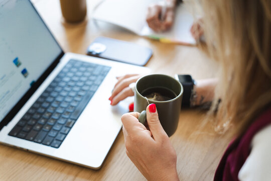 A woman's hands hold a mug of tea while working on a laptop, with a phone and notebook on the desk.