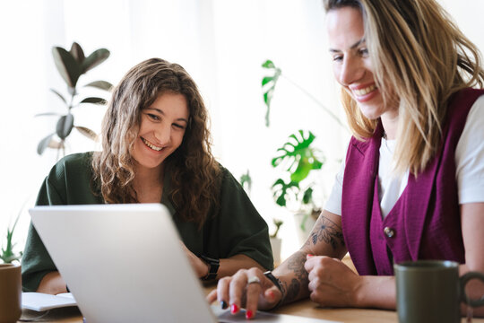 Two women collaborate on a project, smiling as they work together on a laptop at a wooden desk with plants in the background.