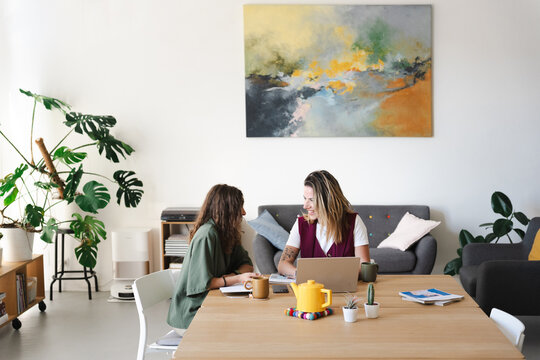 Two women collaborate at a table in a bright, modern office space, discussing work on a laptop.