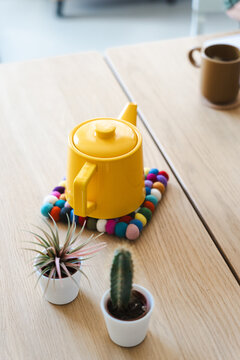 A bright yellow teapot sits on a colorful felt coaster next to two small potted plants on a wooden table.