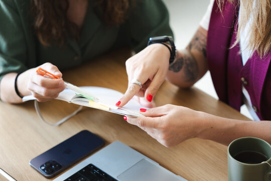 Two women collaborate on a project, reviewing documents and using a laptop and phone on a wooden desk.