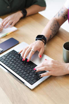 Two individuals collaborate at a wooden desk, one typing on a laptop while the other writes in a notebook, with a smartphone and coffee nearby.