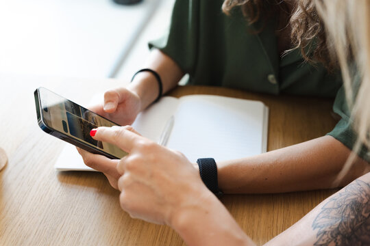 Two women are sitting at a table, one is using her smartphone to browse content while the other looks on.