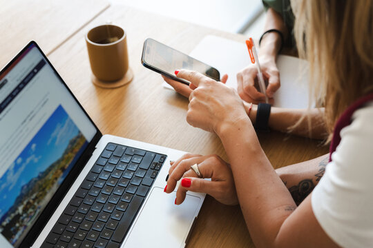 Two women collaborate on a project, one using a laptop and the other a smartphone, while another person takes notes with a pen.
