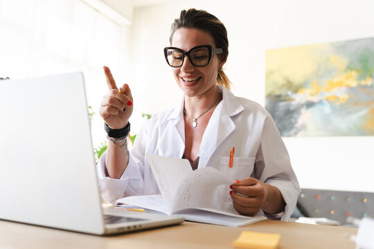 A woman in a lab coat and glasses is working at a desk with a laptop and papers, gesturing with her finger as she speaks.