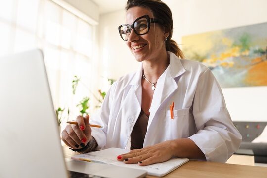 A smiling woman in a lab coat works at a desk with a laptop and notebook, holding a pencil.