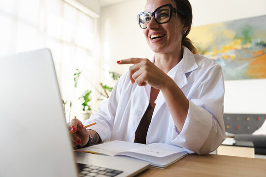 A woman in a lab coat smiles and points while working on her laptop and taking notes in a notebook.