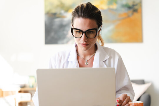 A woman in a white lab coat and glasses works on her laptop, looking focused.