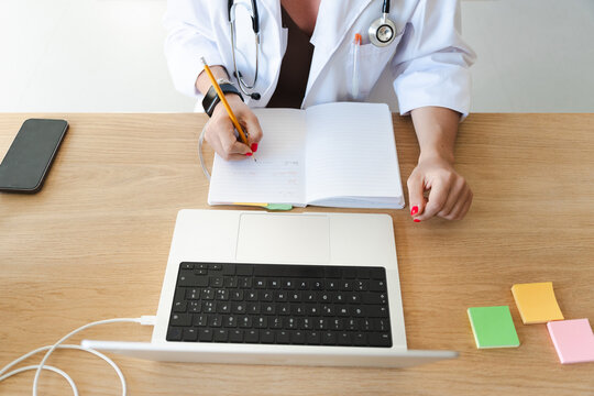 A medical professional in a white coat writes in a notebook at a desk with a laptop, phone, and sticky notes.