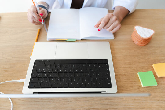 A person in a white coat is writing in a notebook next to a laptop and sticky notes on a wooden desk.