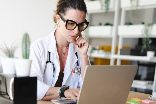 A focused medical professional wearing glasses and a lab coat works on a laptop, with a stethoscope around her neck.