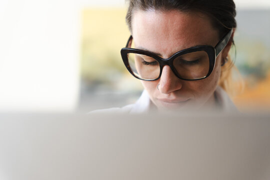 A woman wearing glasses looks down at a laptop screen, concentrating on her work.