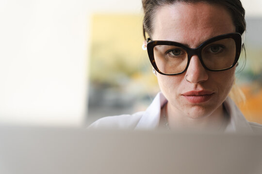 A woman wearing glasses looks intently at a laptop screen, her face illuminated by the device's glow.