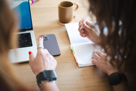 Two people are working together at a desk, one using a laptop and the other writing in a notebook, with a phone and coffee cup nearby.