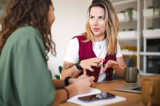 Two women are engaged in a discussion at a table, one taking notes while the other gestures with her hands.