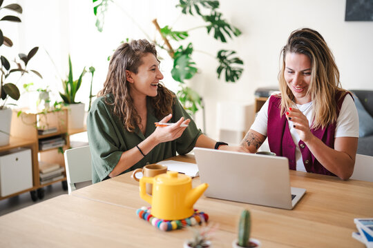 Two women collaborate at a table with a laptop, mugs, and plants, engaged in discussion and smiling.
