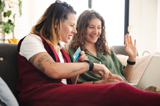 Two women are sitting on a couch, one holding a credit card and looking at a laptop while the other smiles and waves.