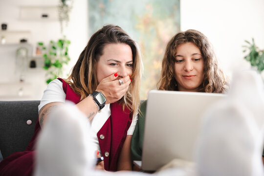 Two women are sitting on a couch, looking at a laptop. One woman covers her mouth with her hand, showing surprise or shock.