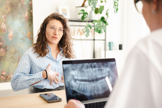 A woman with glasses discusses a dental x-ray on a laptop with a professional in a bright, modern office setting.