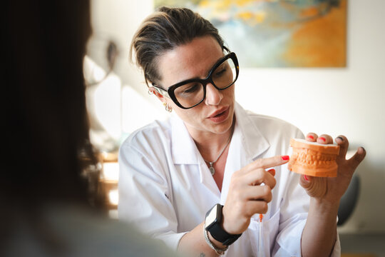 A dentist in a white coat and glasses explains dental anatomy using a model of teeth to a patient in a consultation room.