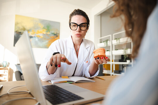 A medical professional in a white coat and glasses explains a dental model to a patient during a consultation.
