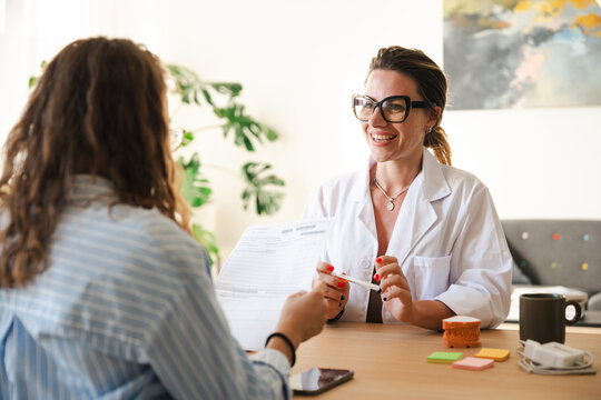 A doctor in a white coat smiles while discussing medical results with a patient, holding a pen and paperwork.
