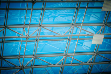 Fototapeta premium Modern architectural ceiling with glass panels and steel framework, showcasing a bright blue sky and natural light filtering through the transparent structure