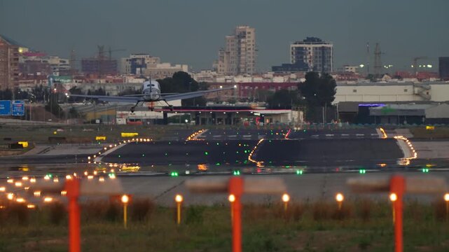 Small airplane on final approach over brightly lit runway threshold at dusk, colorful approach lights and wet tarmac reflections below, city skyline in the background