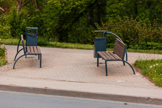 Two metal benches with wooden slats positioned on a paved area beside a walking path, surrounded by greenery and a trash bin nearby