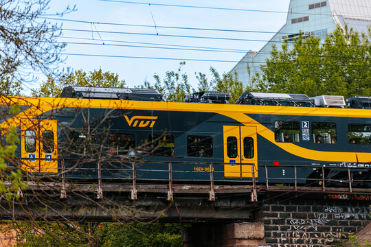 Riga, Latvia - May 15, 2026: Yellow and black train VDV crossing a bridge with green trees and modern building in the background on a clear day