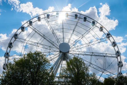 Large Ferris wheel with white cabins against a bright blue sky filled with clouds and sunlight shining through, surrounded by green trees in a public park setting