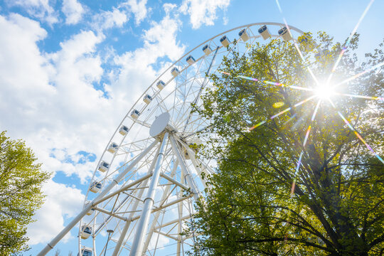 Large white ferris wheel towering above green trees under a bright blue sky with fluffy clouds and sunlight creating a starburst effect in the background