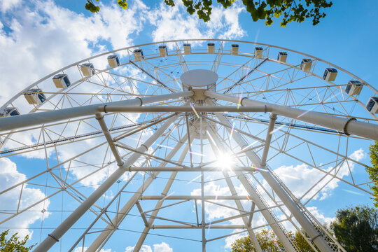 Large white Ferris wheel viewed from below against a bright blue sky with fluffy clouds and sunlight shining through the structure in a park setting