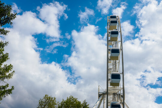 Large white Ferris wheel with blue cabins against a bright sky filled with fluffy clouds and green trees in the foreground during daytime