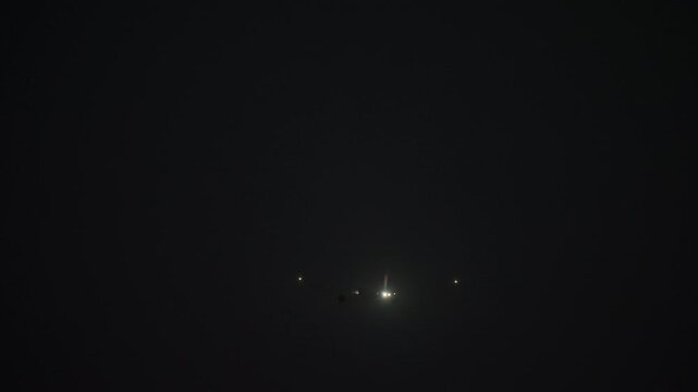 Commercial airplane crosses pitch-dark night sky visible only by red beacon and white strobe lights, wings barely silhouetted against complete darkness