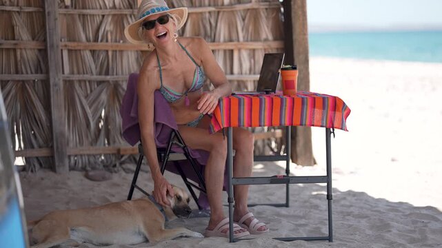 Portrait of mature woman using computer, and phone on tropical beach during vacation with her dog nearby.