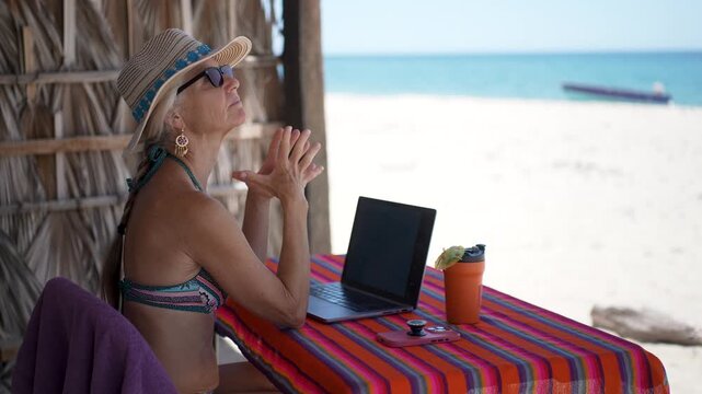 Mature woman uses computer and phone on beach while on vacation. She is thinking for solutions while working in a tropical setting.