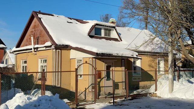 Snow covered cottage under clear blue sky, sunlit roof and chimney, fenced yard with snow piles, leafless birch tree casting long.