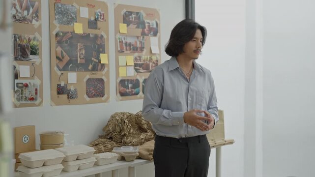 Office worker bright indoor room, side view of person table with packaged food and paper boxes, adult man standing creative studio with wall collage and bulletin board, thoughtful planning scene