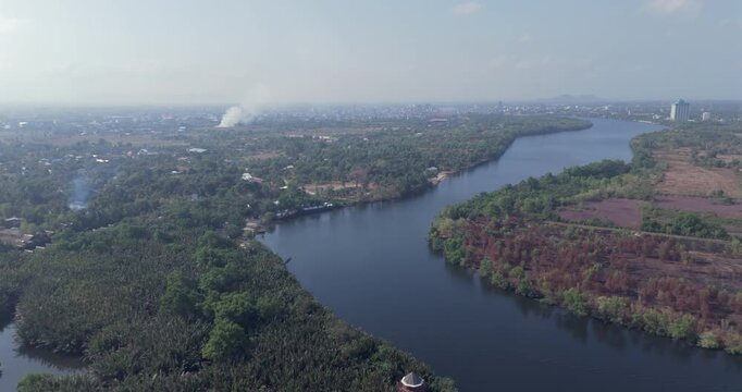 Backward aerial view of a winding river flanked by tropical palm forests and fields, leading toward the hazy town of Kampot with distant smoke on the horizon