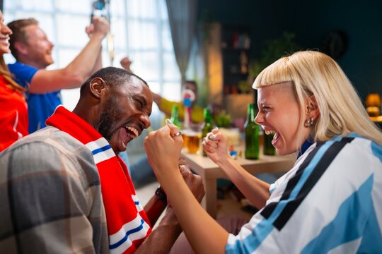 Group of friends watch a sports event at home. They cheer, laugh, and show excitement. Drinks and snacks are on the table. Everyone enjoys the moment together.