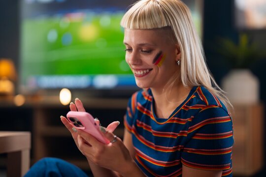 A woman sits on the floor, looking at her phone while watching a sports game on TV. She has face paint and a striped shirt. The room has light from the TV in the background.