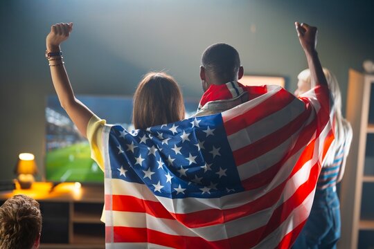 Friends gather in a living room to watch a soccer match on TV. Two people wear an American flag, cheering as they enjoy the game together. It is an evening event filled with excitement.