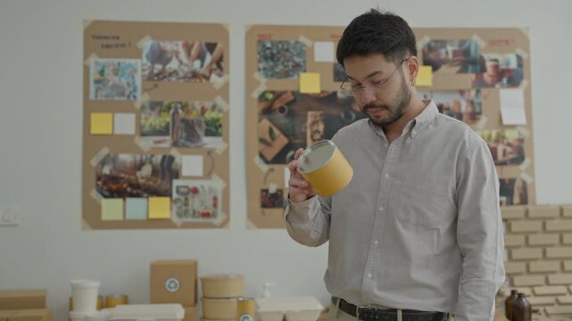 Man holding yellow cup office workspace with bulletin board, project notes, desk, shirt, and businessman holding paper tape with packaging samples, planning board, cardboard tube, thoughtful