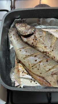 Two sea bass roasted in oven on black pan, close-up showing golden seasoned fillets on aluminum foil atop gas stove, wooden counter.